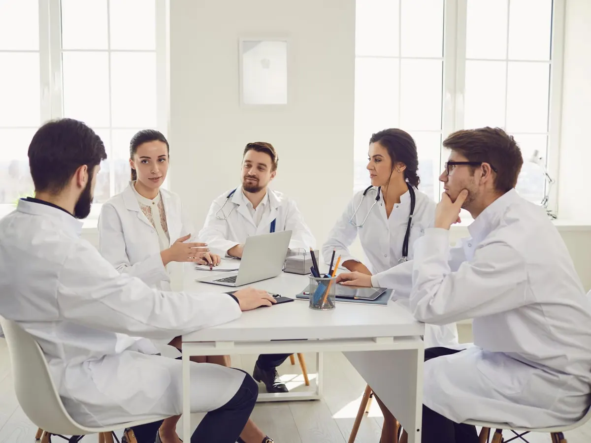 Stock photograph of models: five doctors in white coats talk around a table in a bright office