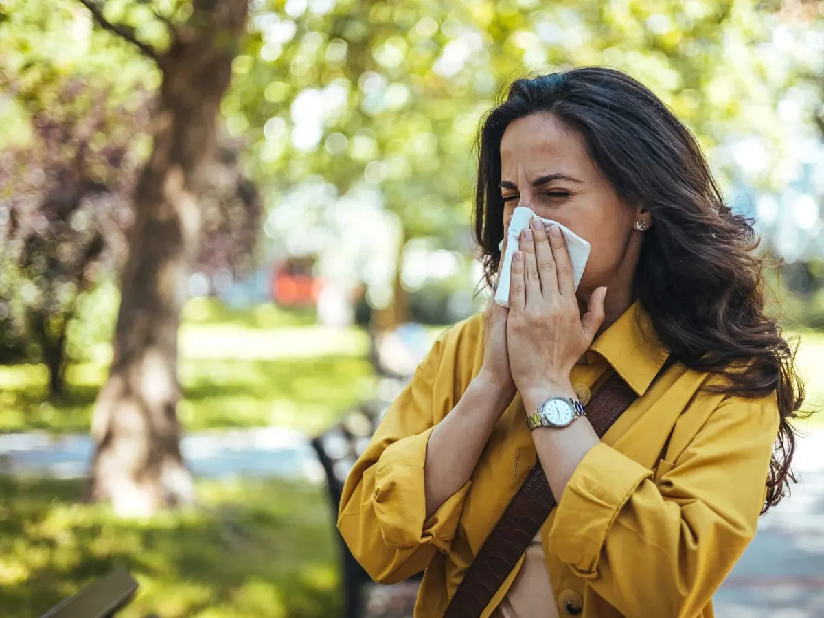 Stock photograph of a model: a woman in a yellow jacket sneezes into a tissue in a sunlit park