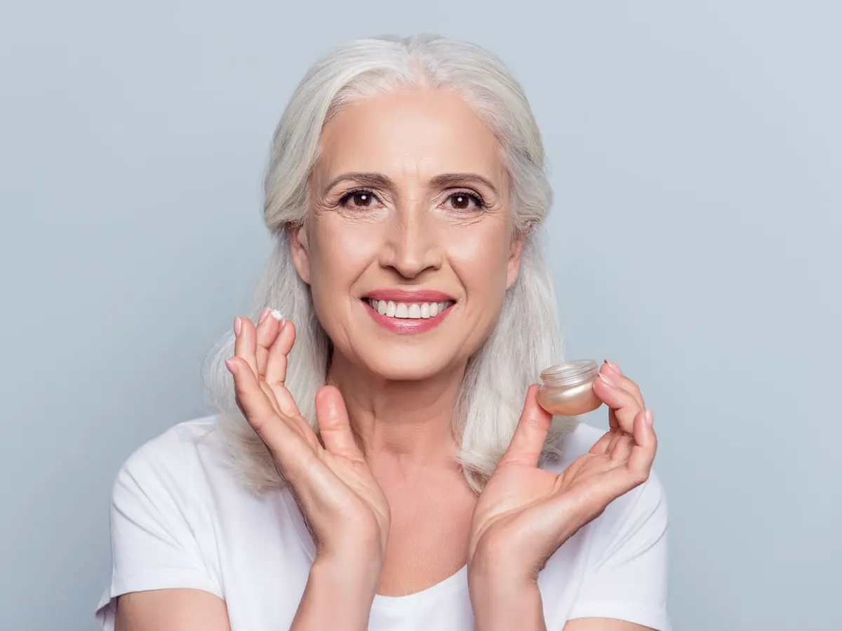 Stock photograph of a model: a smiling grey-haired woman holds a jar of face cream and a dab of it on her fingertip