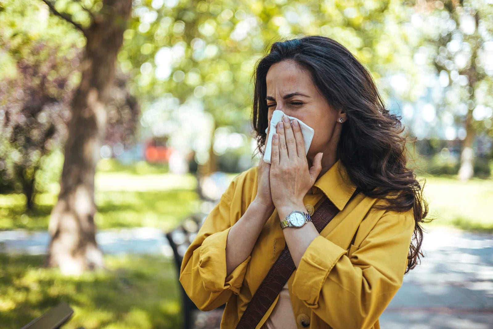 Stock photograph of a model: a woman in a yellow jacket sneezes into a tissue in a sunlit park