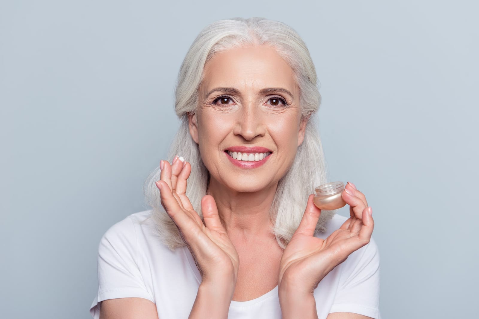 Stock photograph of a model: a smiling grey-haired woman holds a jar of face cream and a dab of it on her fingertip