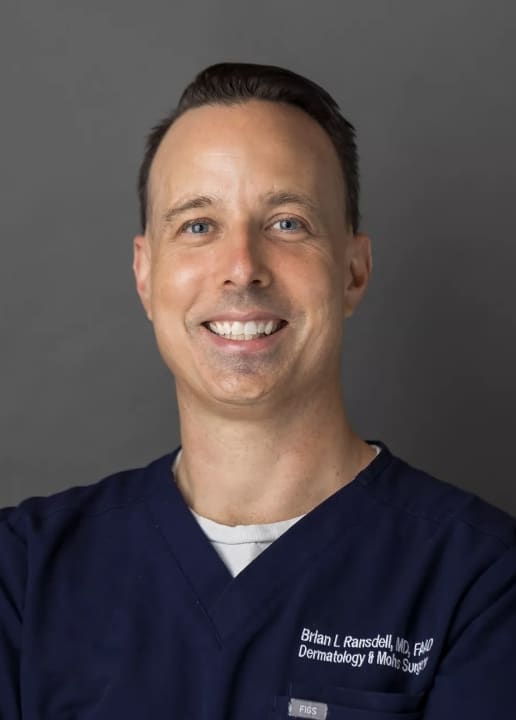 Portrait of Dr. Brian Ransdell, MD, FAAD, smiling, in navy scrubs